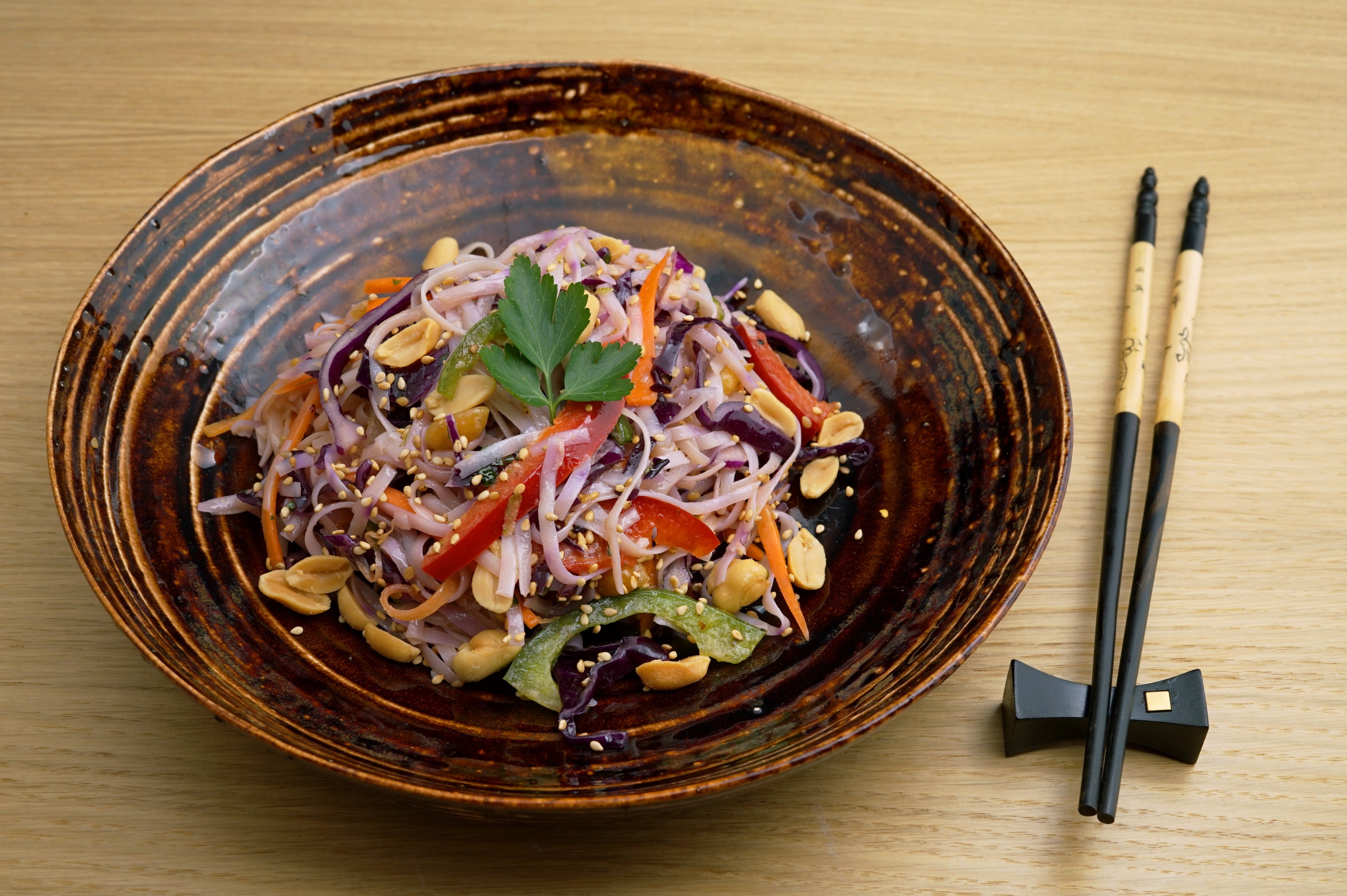 Bunte Schale mit Reisnudel-Gemüse-Salat, garniert mit Erdnüssen und Sesam. Essstäbchen liegen neben der Schale auf einem Holzuntergrund. Frisches Petersilienblatt verziert den Salat.