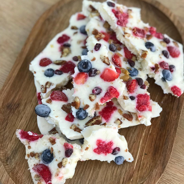 Joghurt Bites sind eine selbstgemachte Köstlichkeit aus Naturjoghurt, Nüssen und roten Früchten die gefroren sind. Sie sehen aus wie zerbrochene Teile einer Platte und wurden auf einem Holztablett serviert.